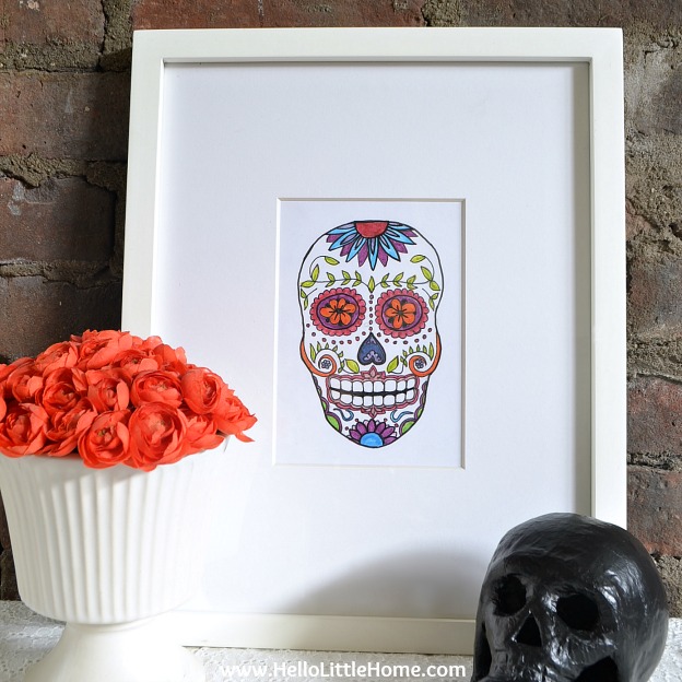 A framed print, flowers, and skull on a table in front of a brick wall.