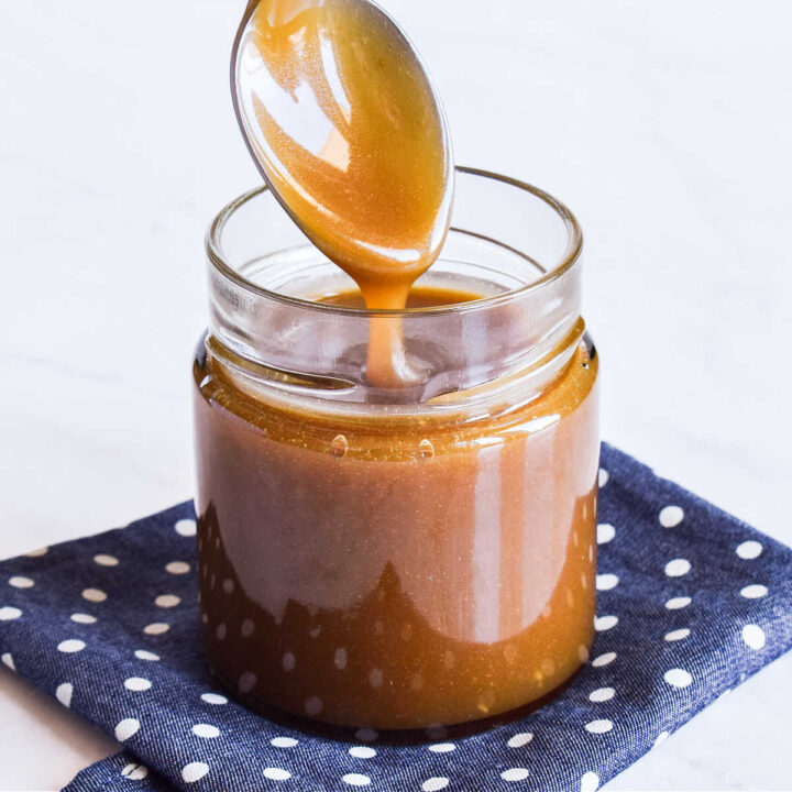 A jar of Salted Caramel Sauce on a polka dot napkin with a spoon drizzling the sauce into it.