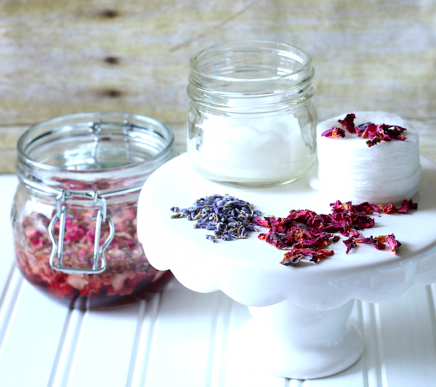 A container of All-Natural Facial Wipes next to dried lavender and rose petals on a white pedestal.