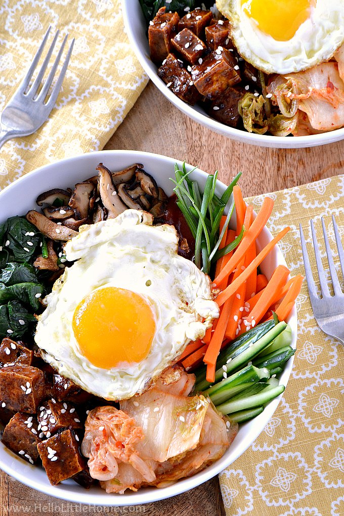 Two bowls of Tofu Bibimbap topped with fried eggs on a table with napkins and forks.