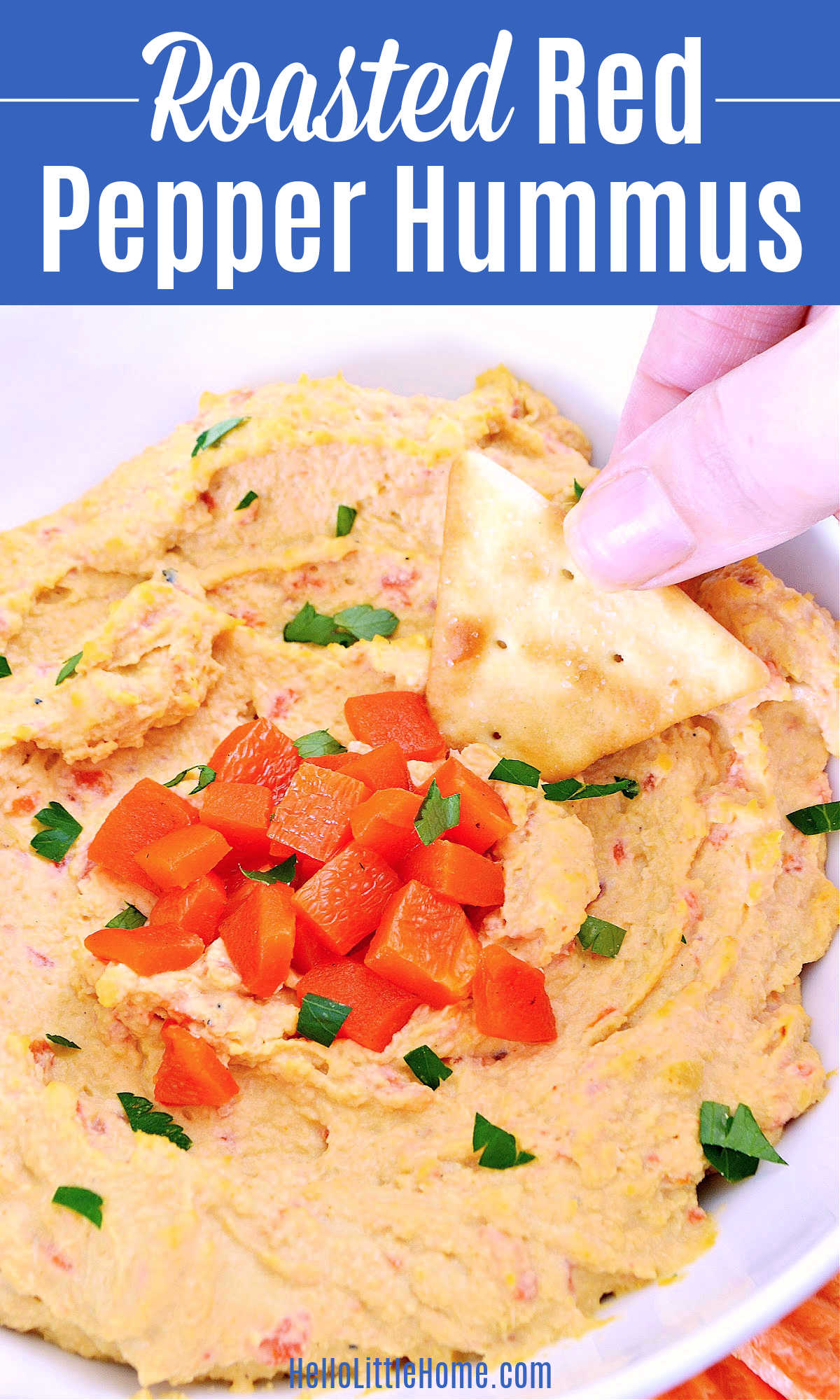 A hand dipping a pita chip in a bowl of Roasted Red Pepper Hummus.
