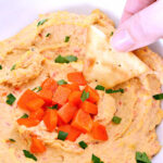 A hand dipping a pita chip in Roasted Red Pepper Hummus.