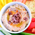 A bowl of olive hummus surrounded by veggies and crackers.