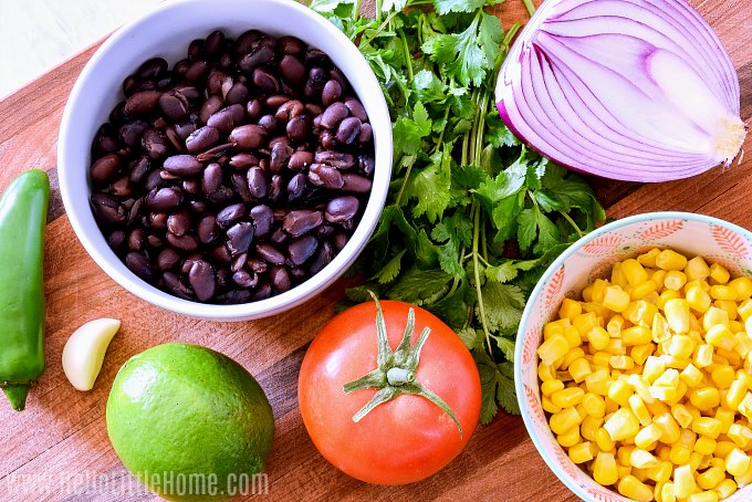 Black Bean Salsa ingredients on a wood board: beans, jalapeno, garlic, lime, tomato, corn, onion, and cilantro.