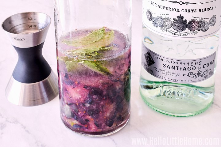 A jigger, glass with rum and muddled blueberries, and rum bottle on a marble counter.