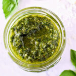 A jar of Pesto Sauce and basil leaves on a marble counter.