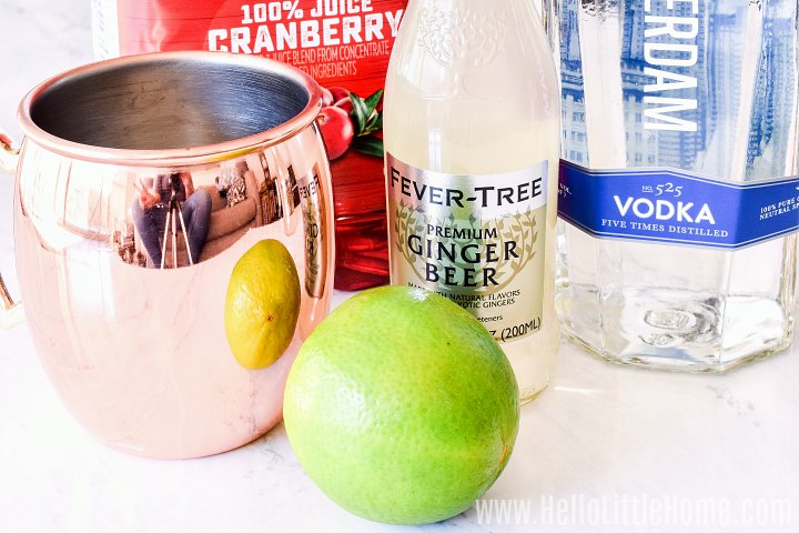 Recipe ingredients on a marble counter: lime, cranberry juice, ginger beer, vodka, and copper mug.