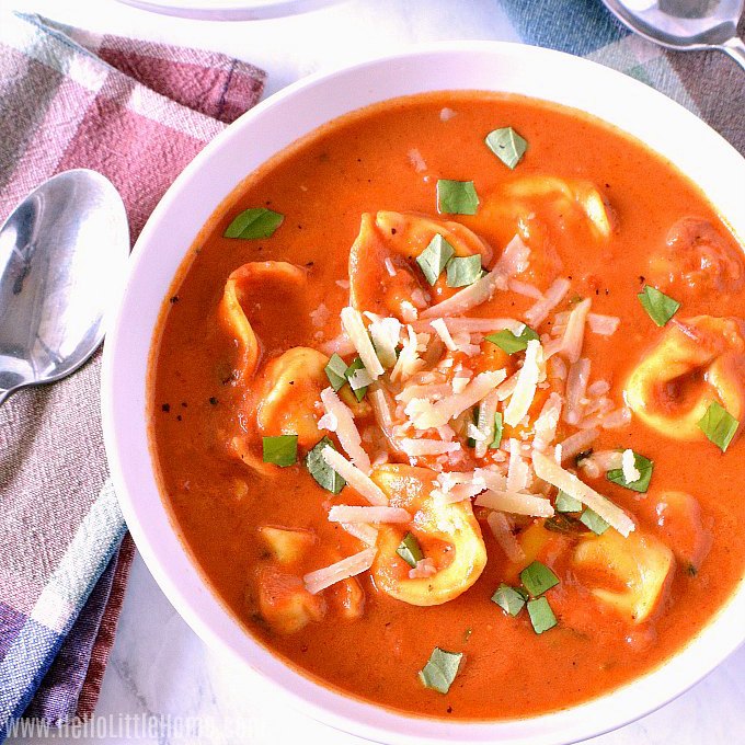 A bowl of tomato tortellini soup next to a plaid napkin and spoon.