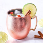 An Apple Cider Mule, cinnamon sticks, and lime slices on a counter.