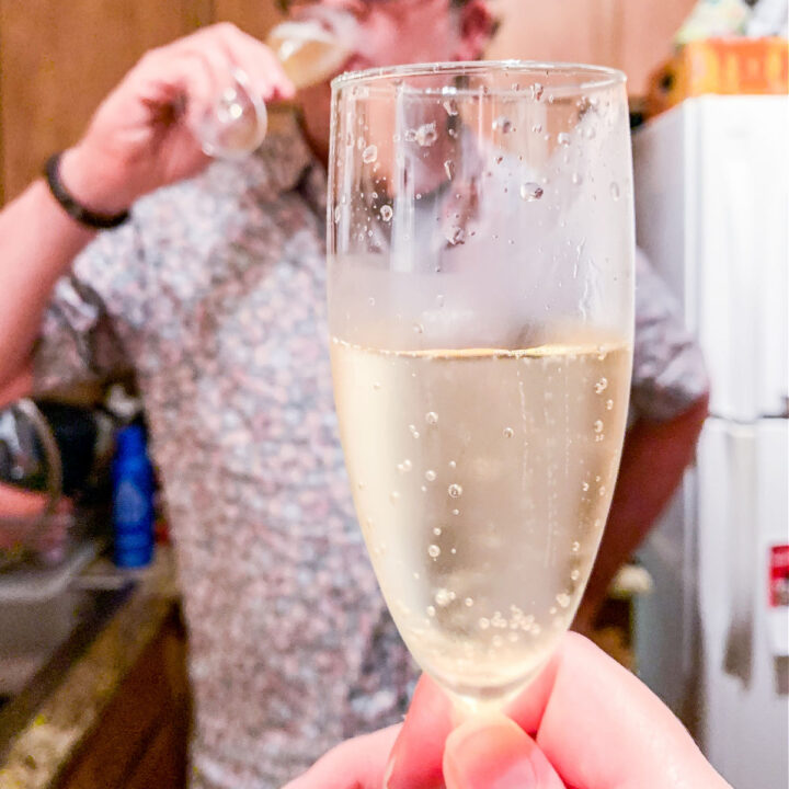 A hand holding a champagne flute in front of a man drinking champagne.