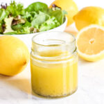 A jar of Lemon Olive Oil Dressing, lemons, and salad greens on a marble counter.