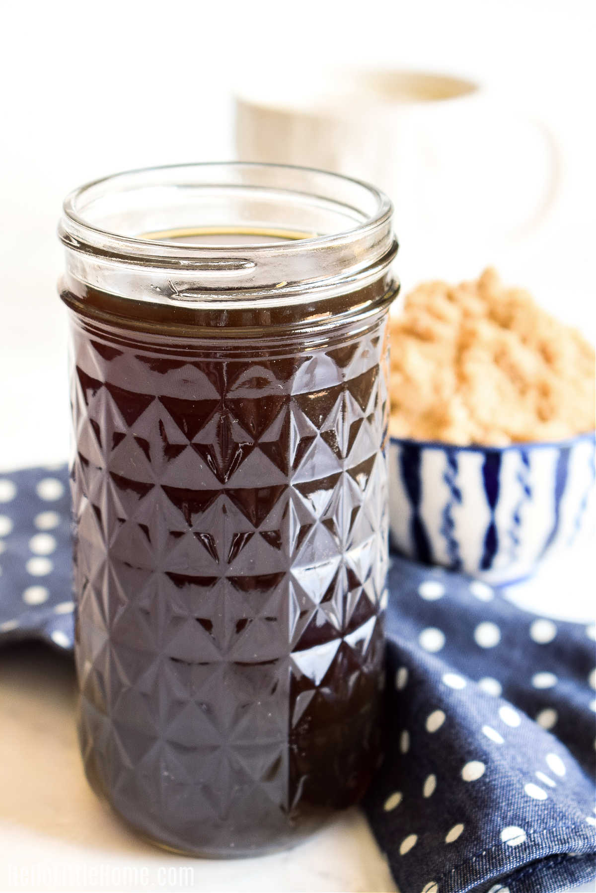 A jar of the finished syrup with a napkin, bowl of brown sugar, and mug in the background.