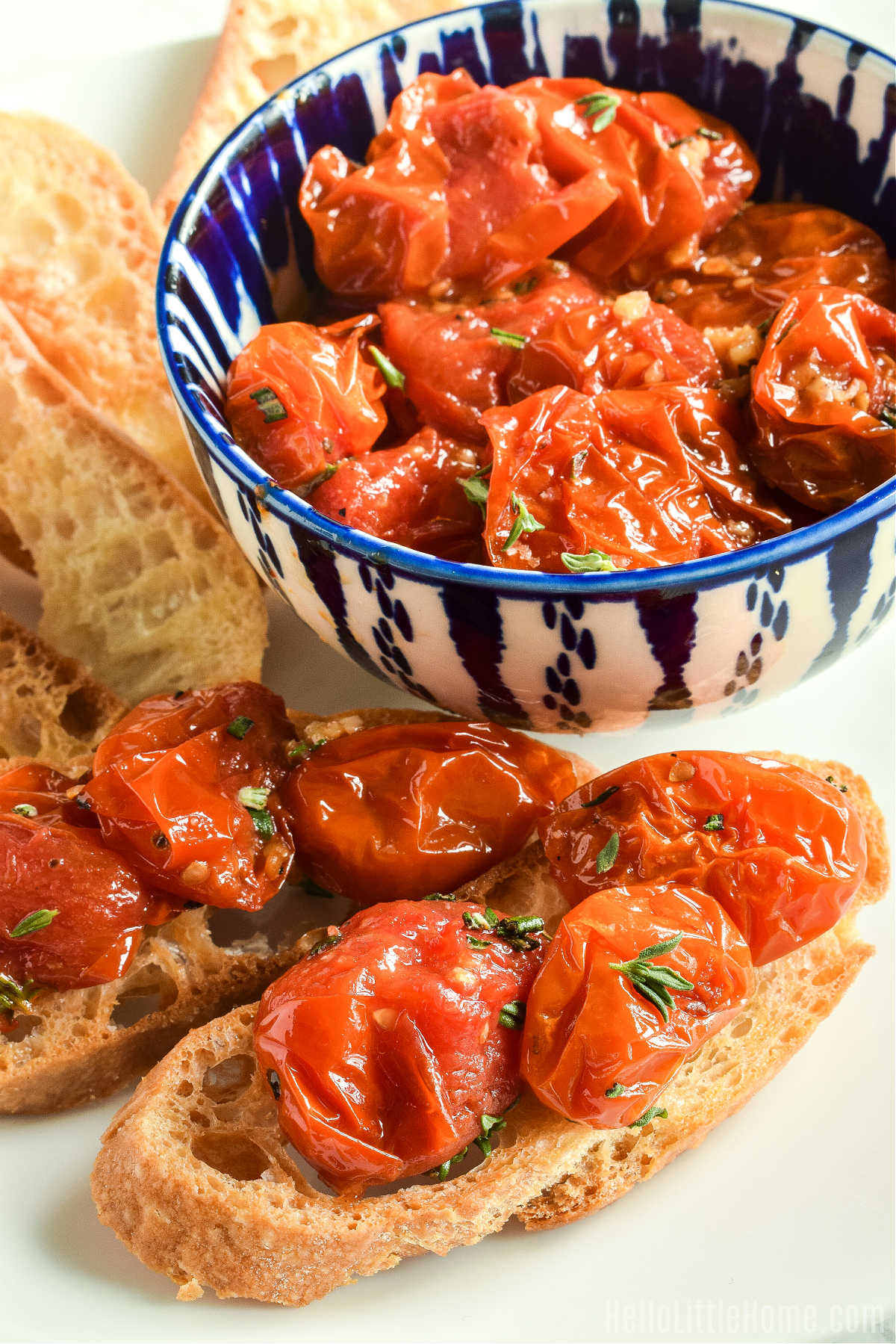 Two toasts topped with the baked tomatoes with a bowl of finished recipe behind them.