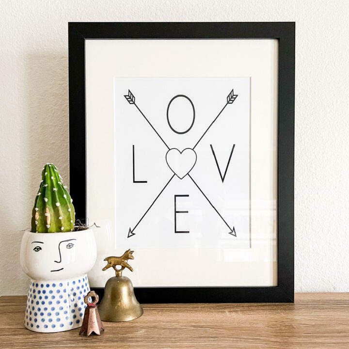 The framed Love Wall Art, a plant, and two small bells on a wood table.