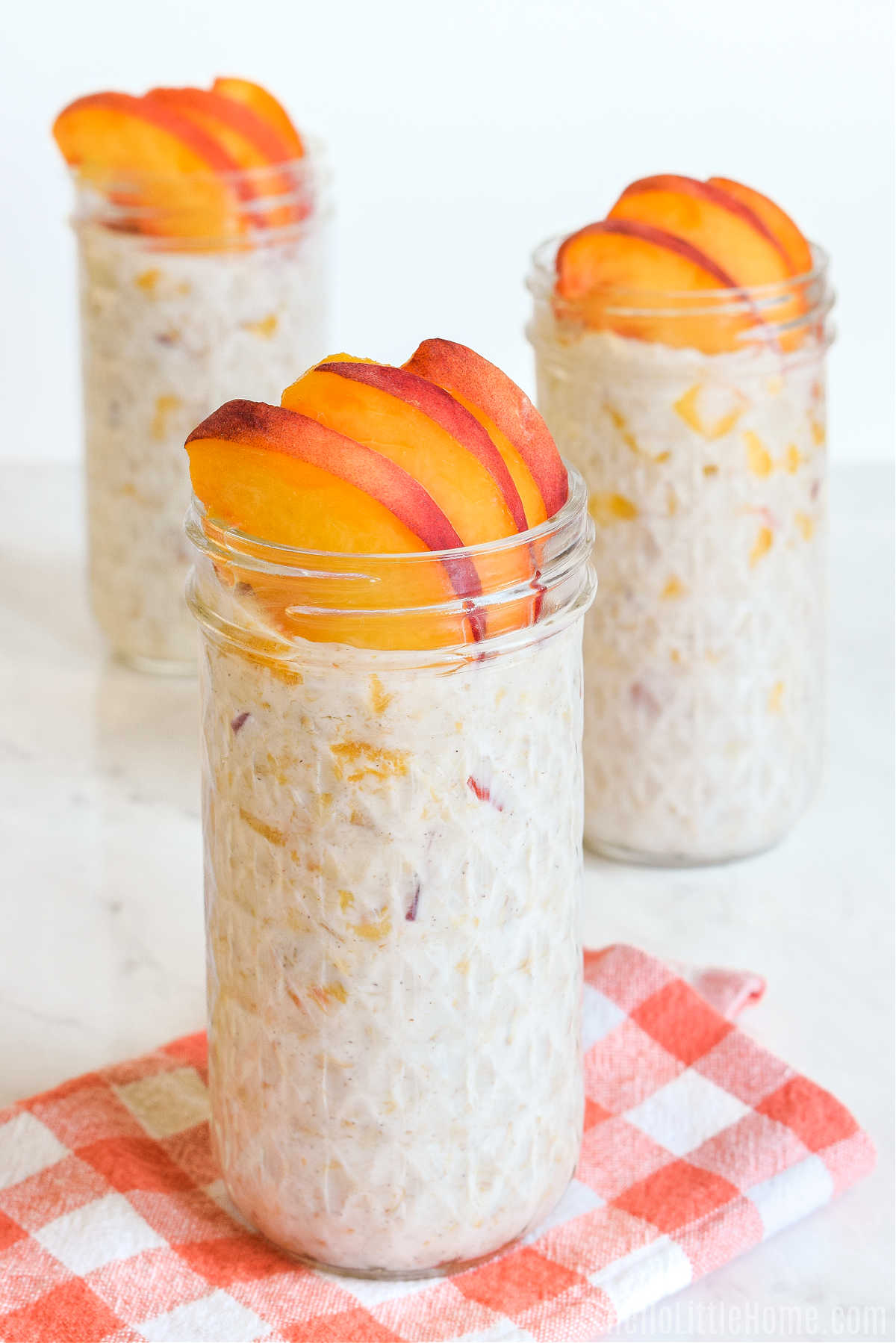Three jars of the peaches and cream oats and a checked napkin on a marble counter.