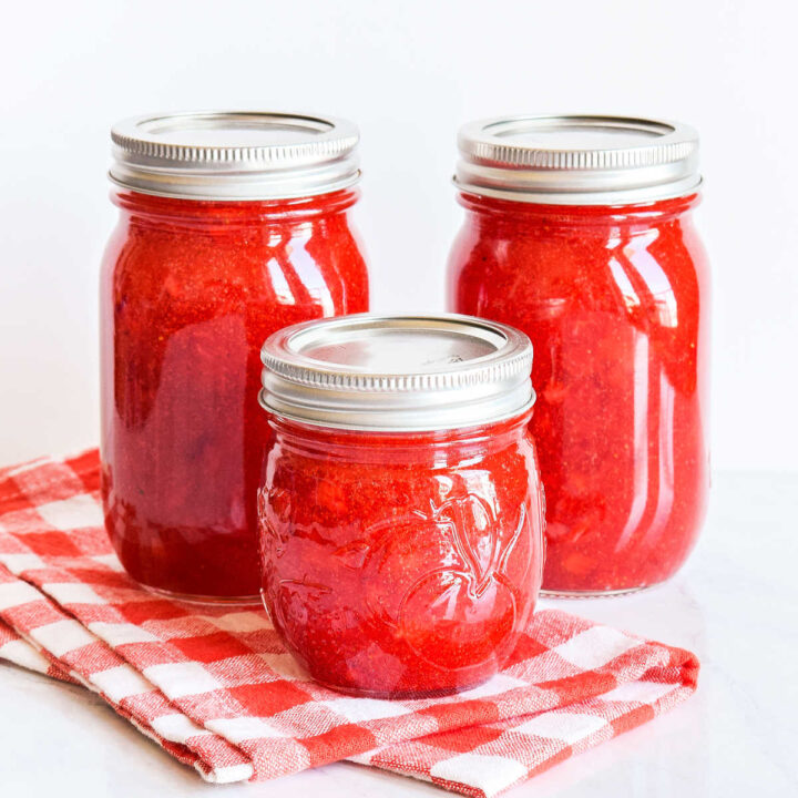Three jars of Strawberry Freezer Jam and a checked napkin on a marble counter.