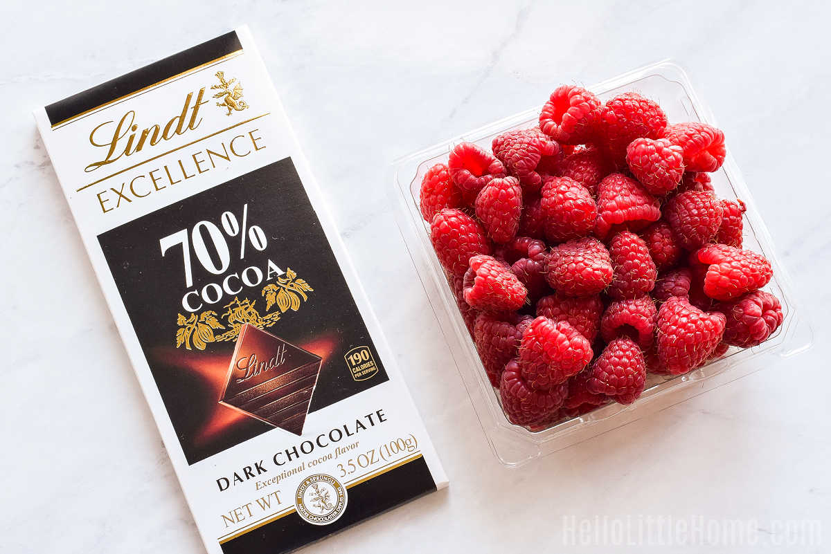 A bar of chocolate and container of raspberries on a white marble counter.