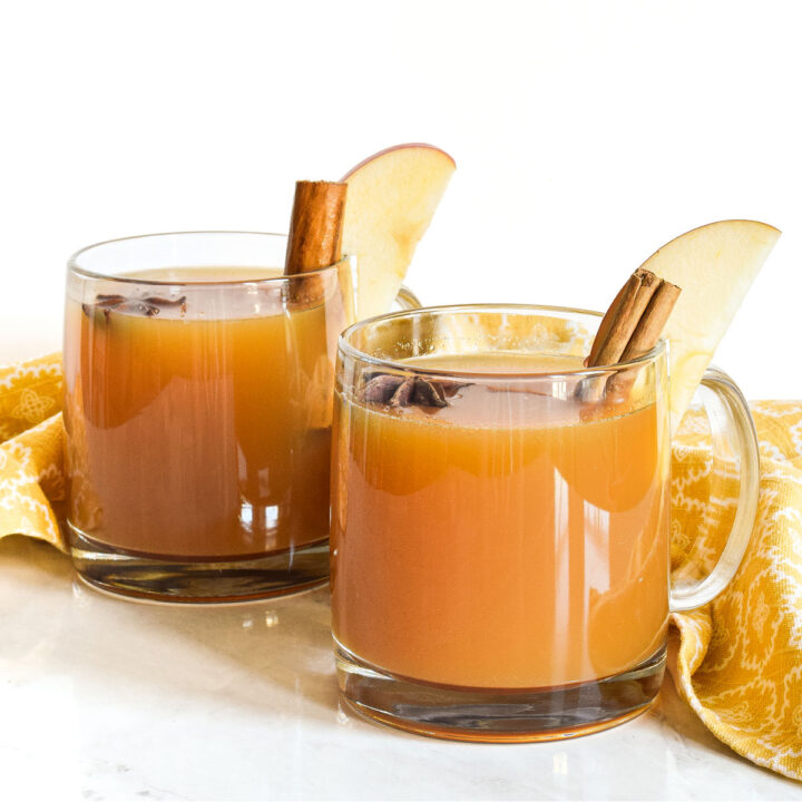 Two mugs of Hot Apple Cider garnished with cinnamon sticks and apple slices.