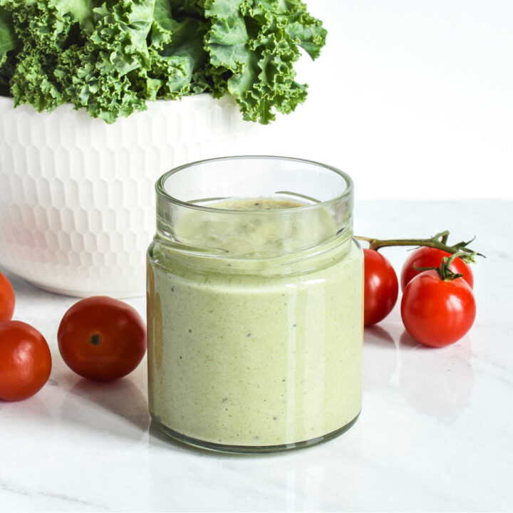 A jar of Pumpkin Seed Salad Dressing, tomatoes, and kale on a marble counter.