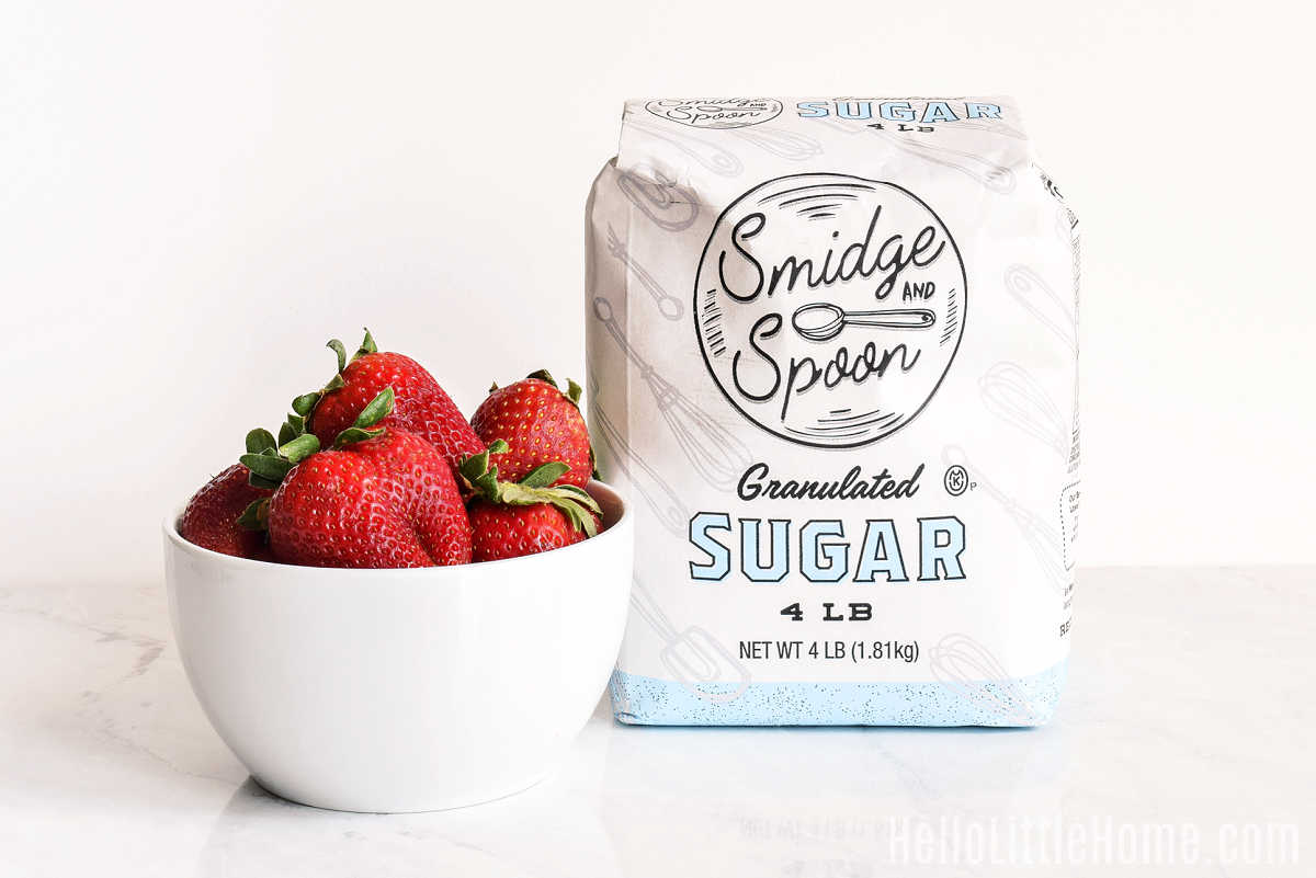 A bowl of strawberries and a bag of sugar on a marble counter.