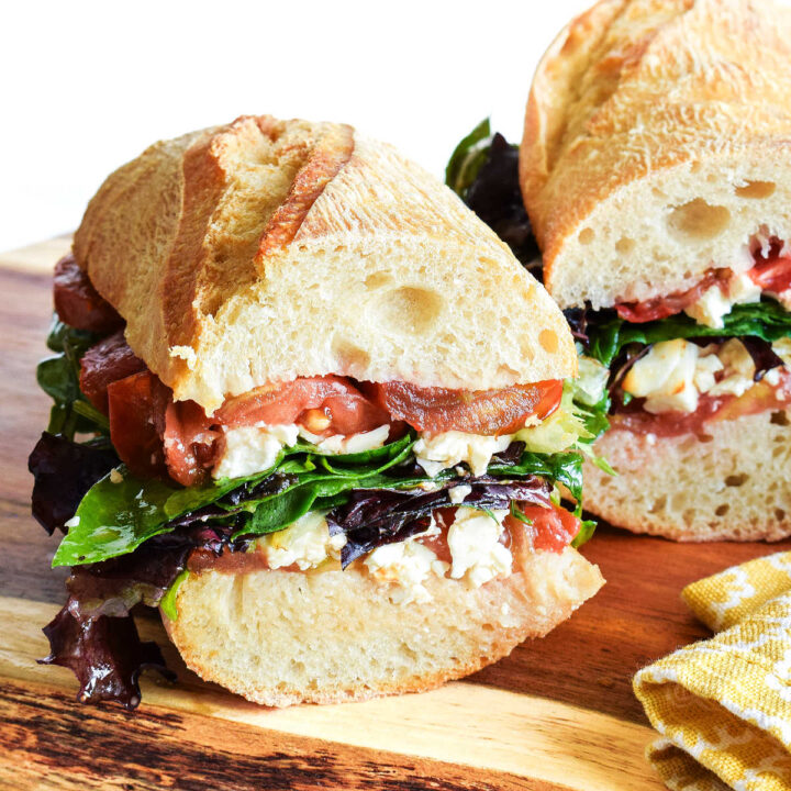 A Feta Sandwich with tomatoes and mixed greens served on a wood cutting board.