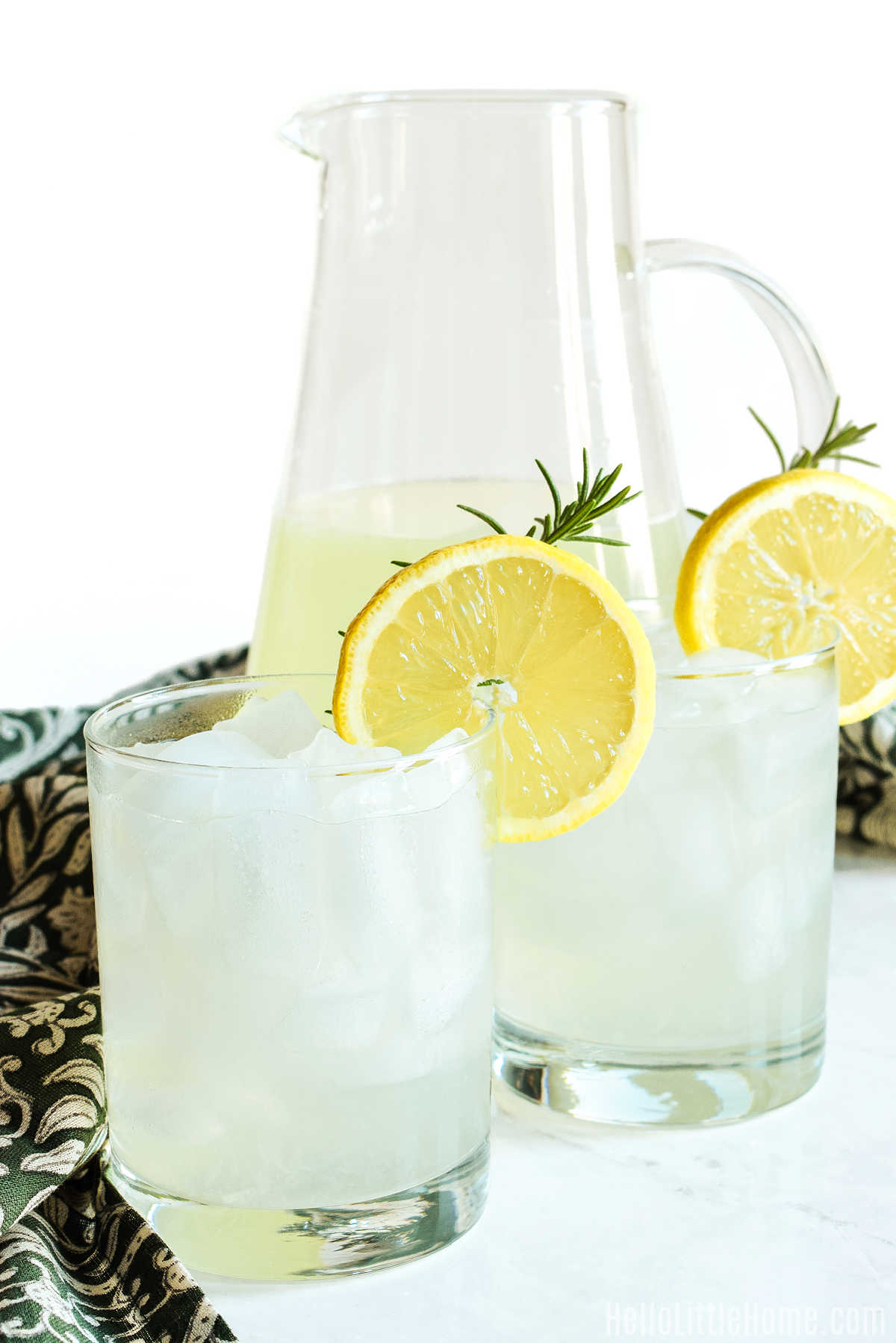 Two glasses of Rosemary Lemonade and a pitcher of the drink on a marble counter.